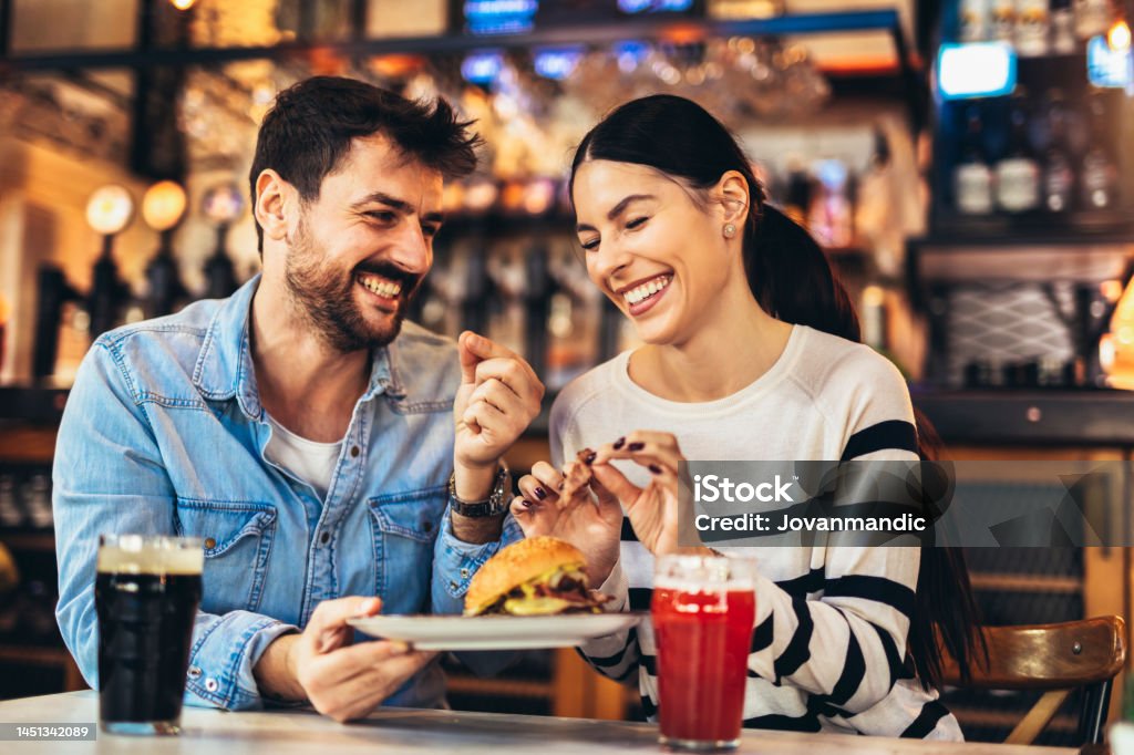 Young couple in love having fun spending leisure time together at restaurant, eating burgers and drinking beer